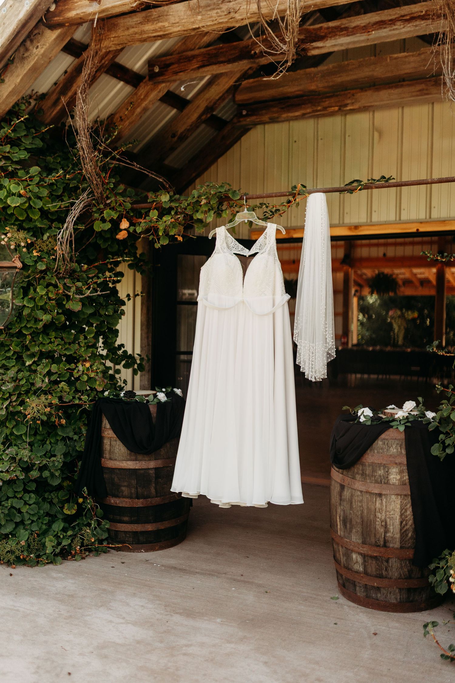 White wedding dress and veil hanging on rustic barn entrance with wooden barrels and ivy.