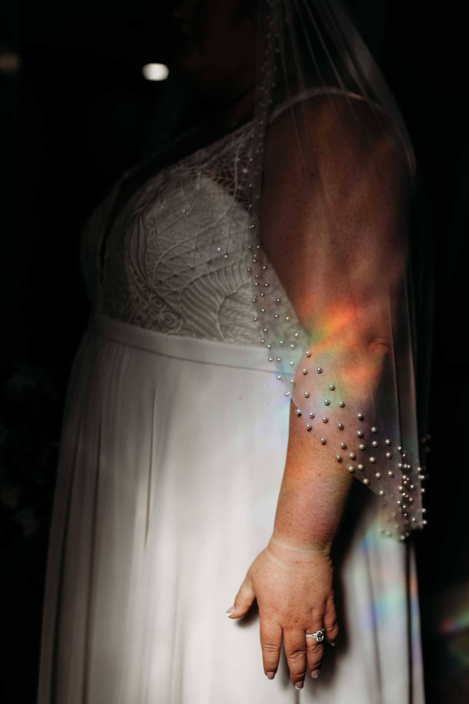 Close-up of bride in pearl-adorned wedding dress sleeve with rainbow light reflection on arm.