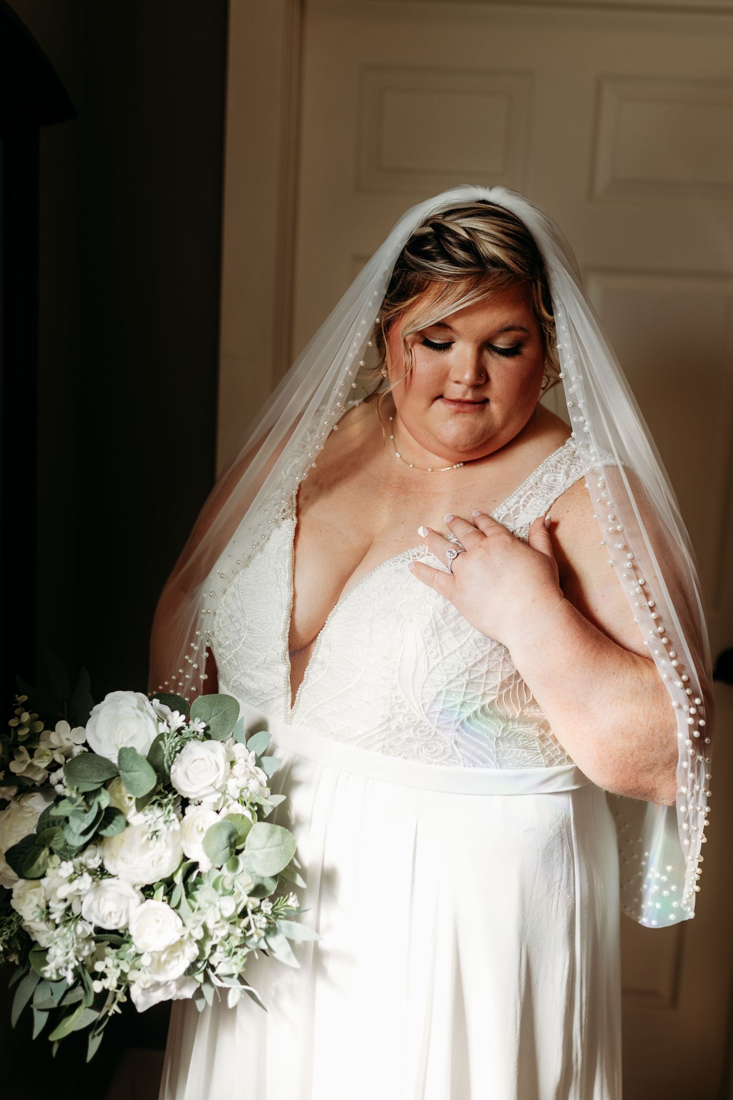 Plus-size bride in lace wedding gown holding white rose bouquet in soft indoor lighting.