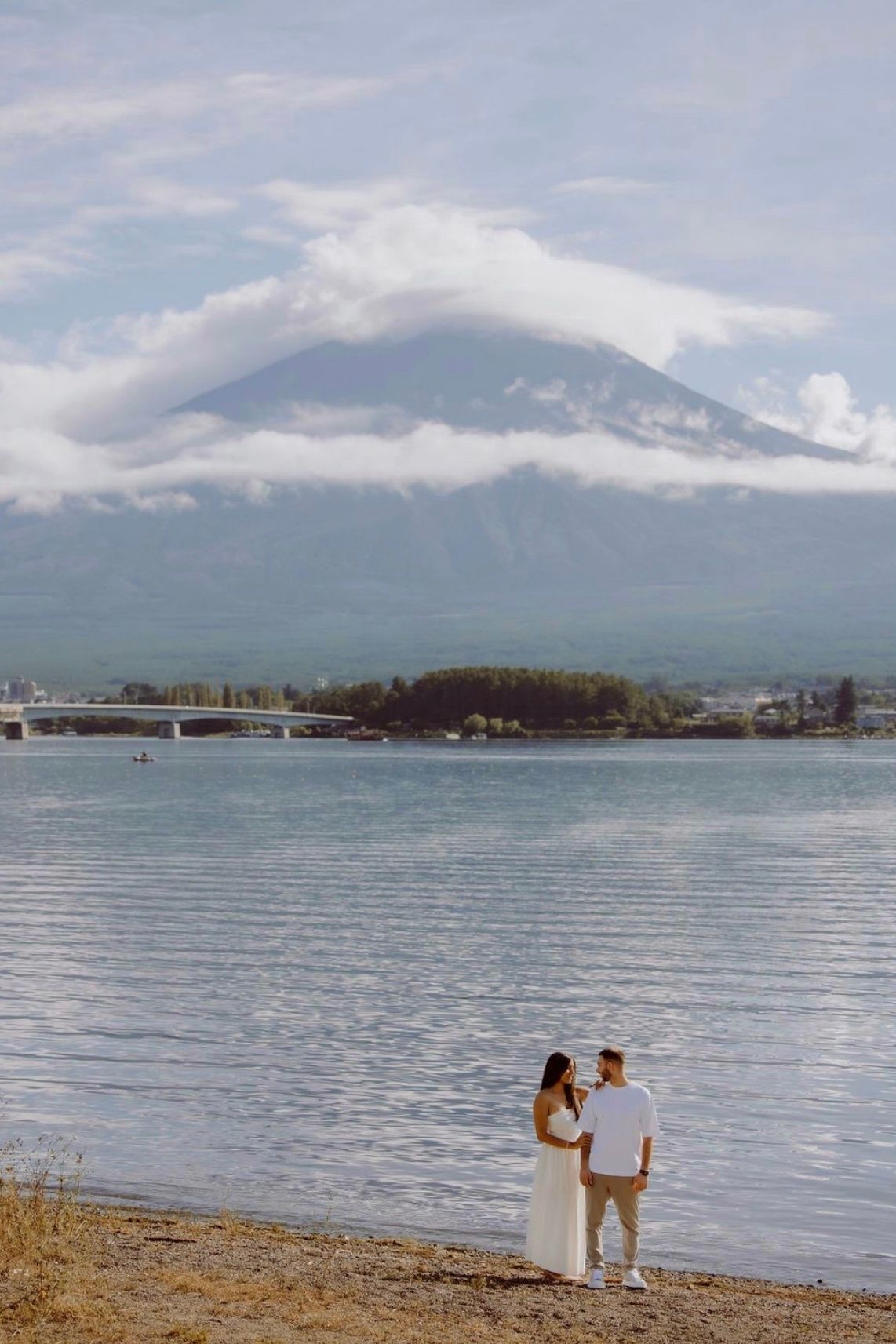 Two figures standing by a lake with Mount Fuji and clouds in the background.
