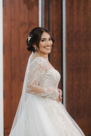 A bride in a detailed lace wedding dress smiles against a wooden backdrop.