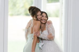 Two women share an emotional embrace, one wearing a white wedding gown and the other in a light blue dress.