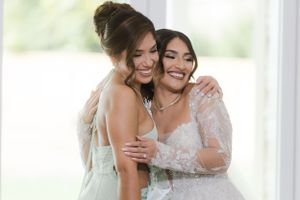 Two women sharing a joyful moment together during a wedding, one in white and one in light blue.