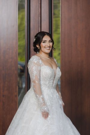 A bride poses in an elegant white lace wedding gown with sheer sleeves against a wooden backdrop.