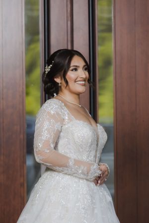 A bride in a stunning lace-sleeved wedding gown poses against wooden doors with a joyful expression.