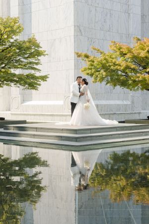 Couple taking wedding photos by a reflective pool surrounded by autumn trees.
