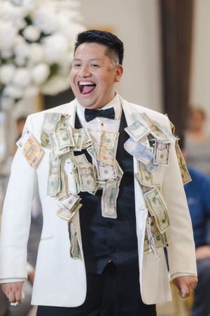 A person in formal attire laughs joyfully with money pinned all over their white vest and jacket.