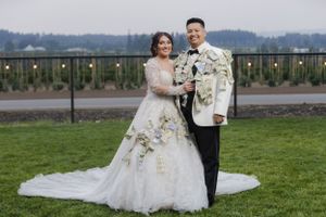 A wedding couple poses outdoors on a grassy field with mountains and string lights visible in the background.