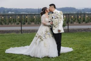 A wedding couple stands together on grass with mountains and string lights creating a romantic backdrop.