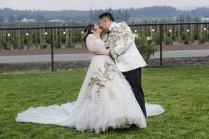 A wedding couple embraces on a grassy field with scenic mountain views and string lights.