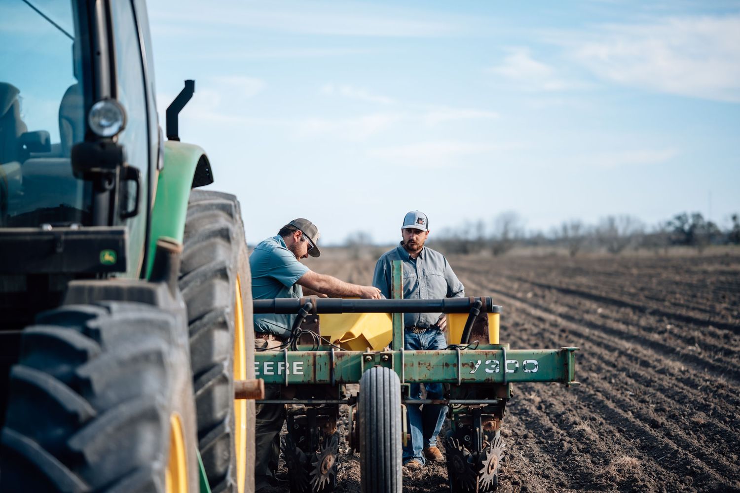 Two farmers working together on planting equipment in a freshly tilled field.