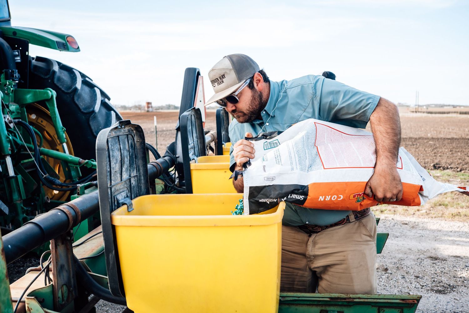 A farmer pours seed or fertilizer into a yellow hopper attached to a green tractor.
