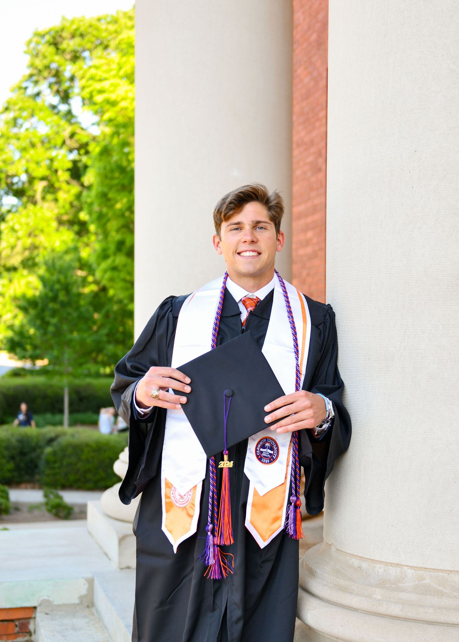 Graduate wearing black cap and gown with honors cords poses by classical column during commencement ceremony.