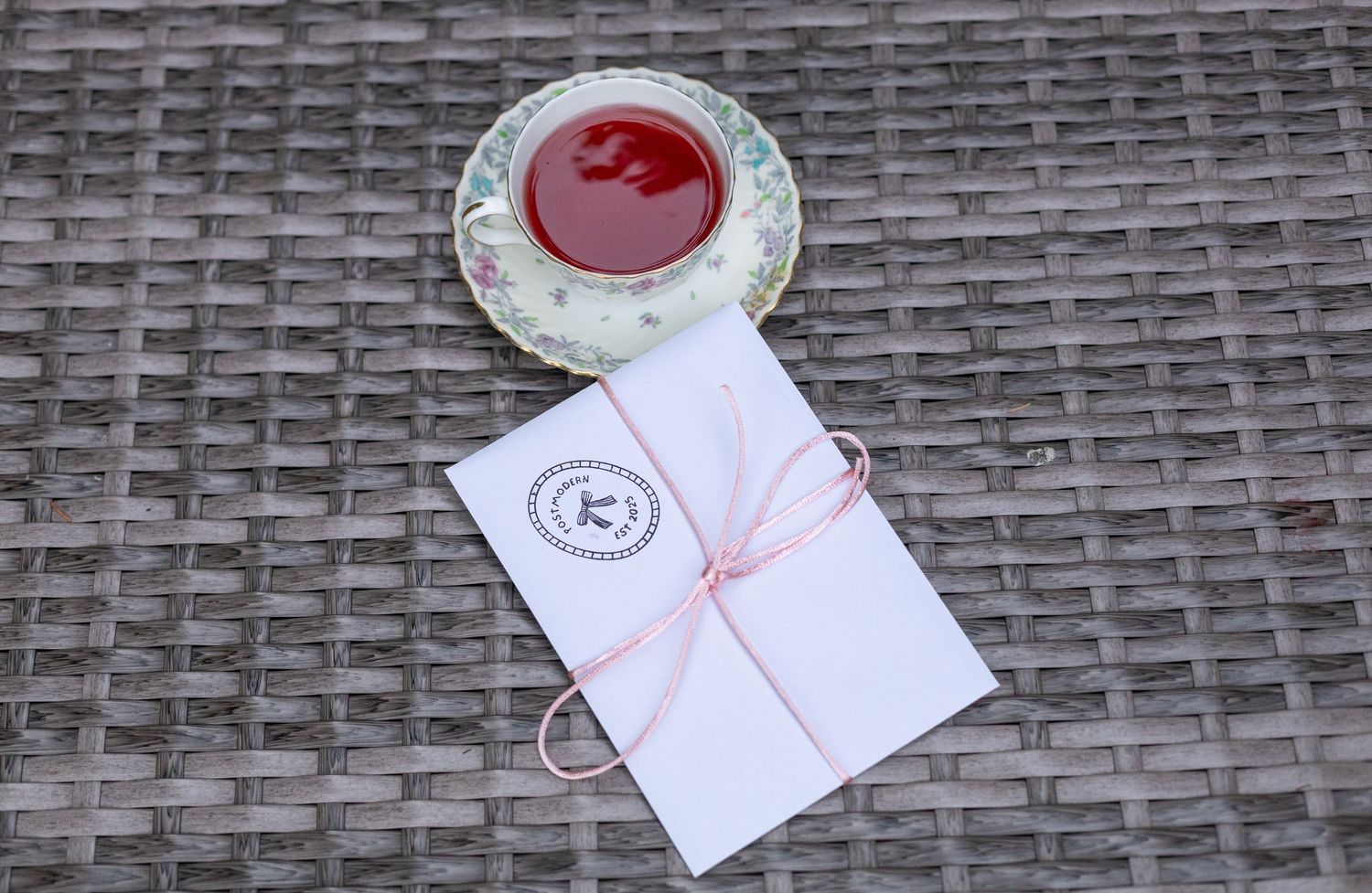 Overhead view of a white envelope with pink string and red drink in decorative teacup on wicker surface.