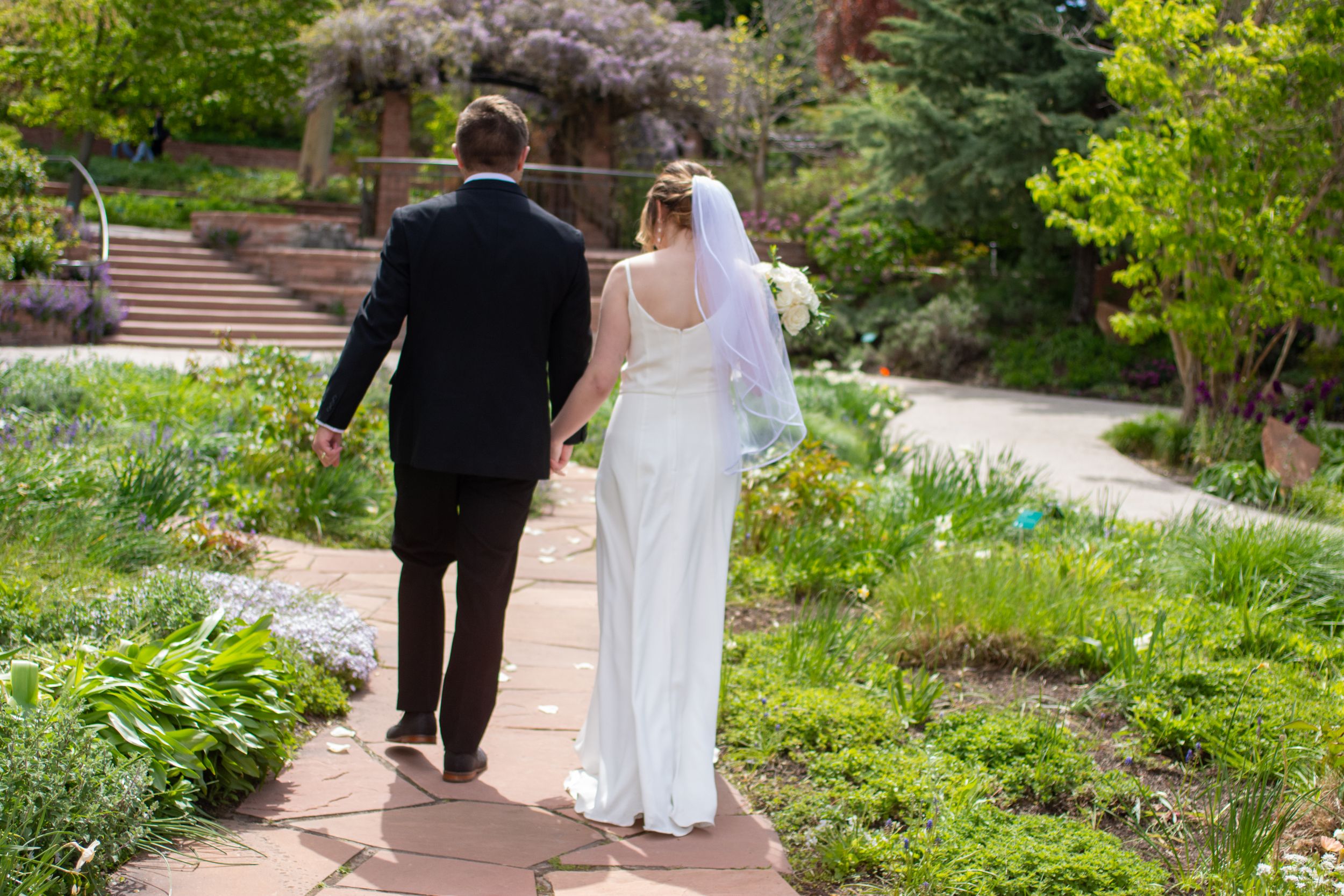 Bride and groom walk hand in hand on wedding day away from camera in a Salt Lake City garden wedding venue.