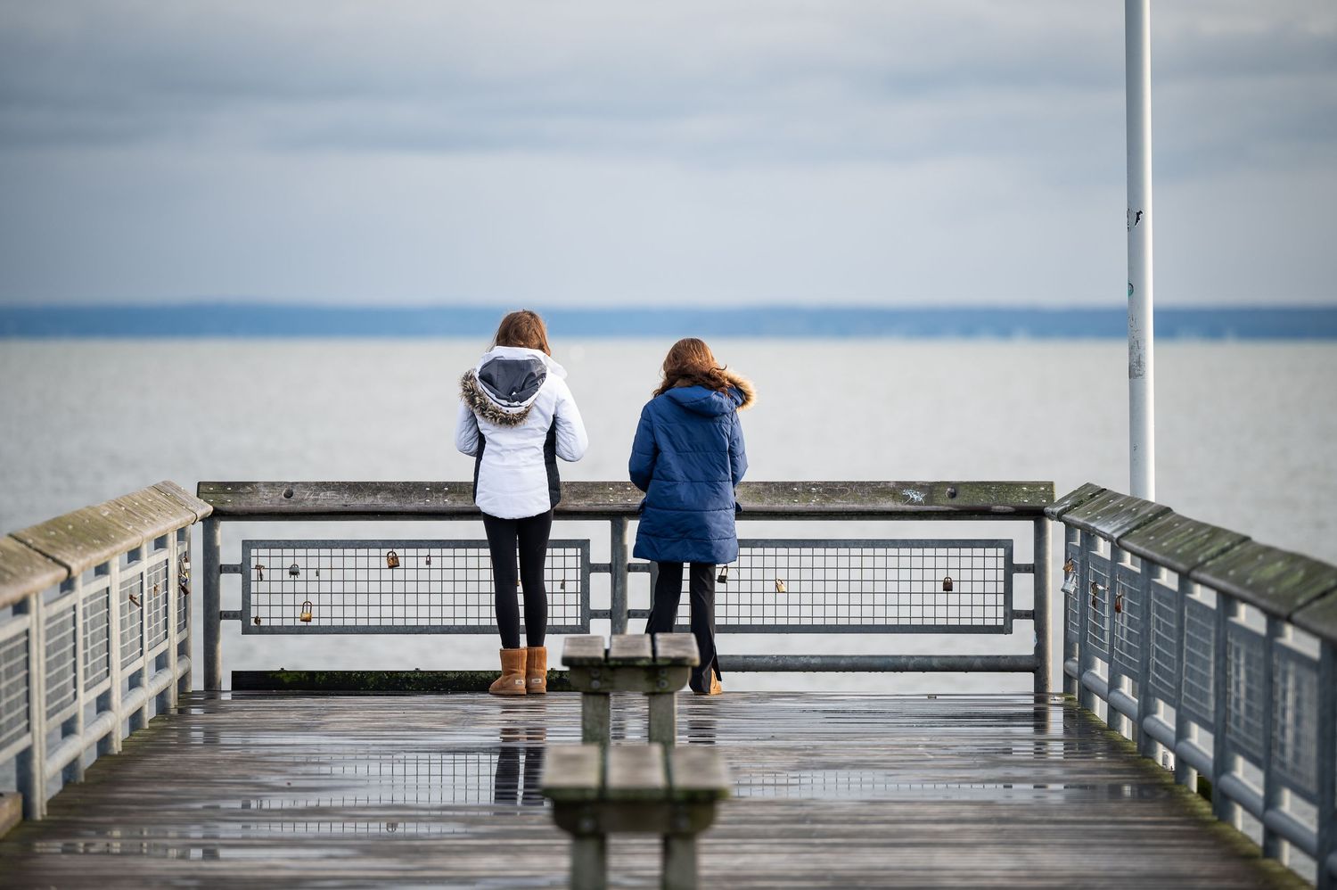 Two people stand at the end of a wet pier overlooking a calm ocean on an overcast day.