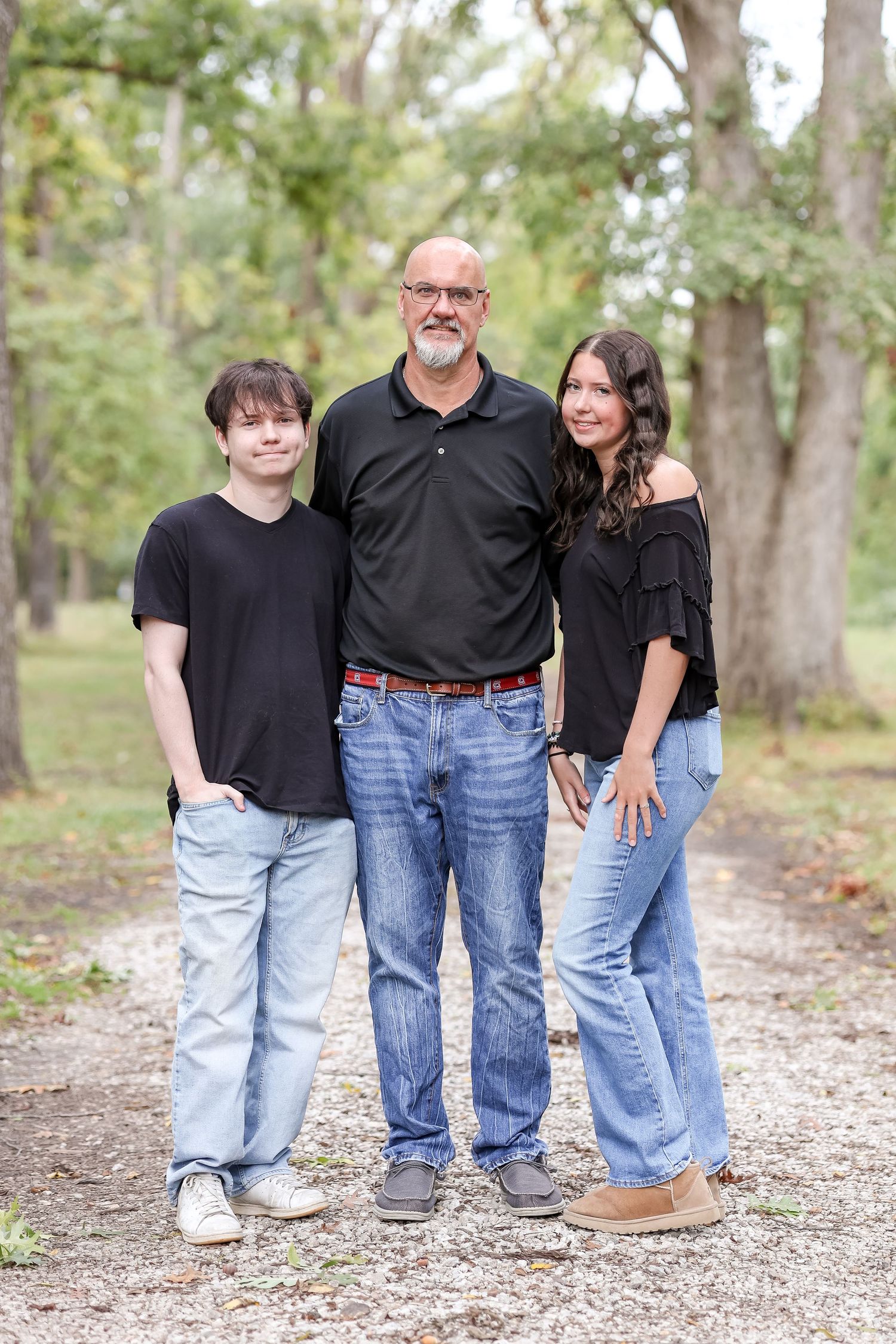 Three people wearing casual outfits stand together on a gravel path in a wooded outdoor setting, all coordinated in black tops.