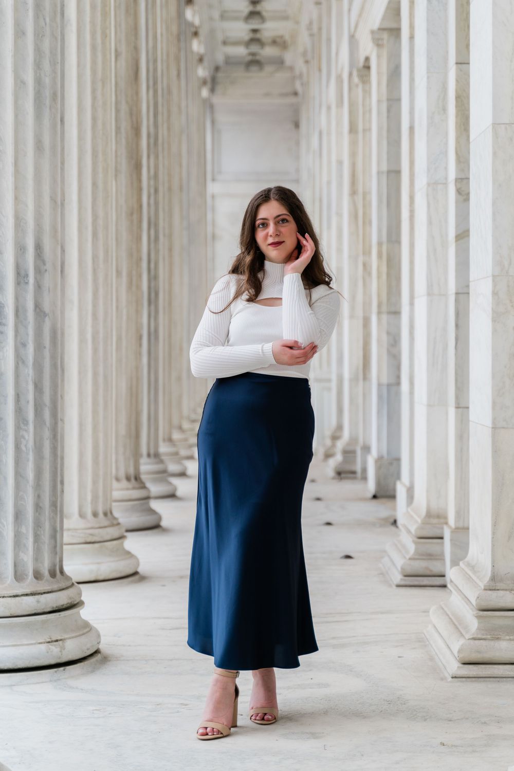 A fashion portrait in a classical colonnade featuring a white turtleneck and navy midi skirt styled with nude heels.