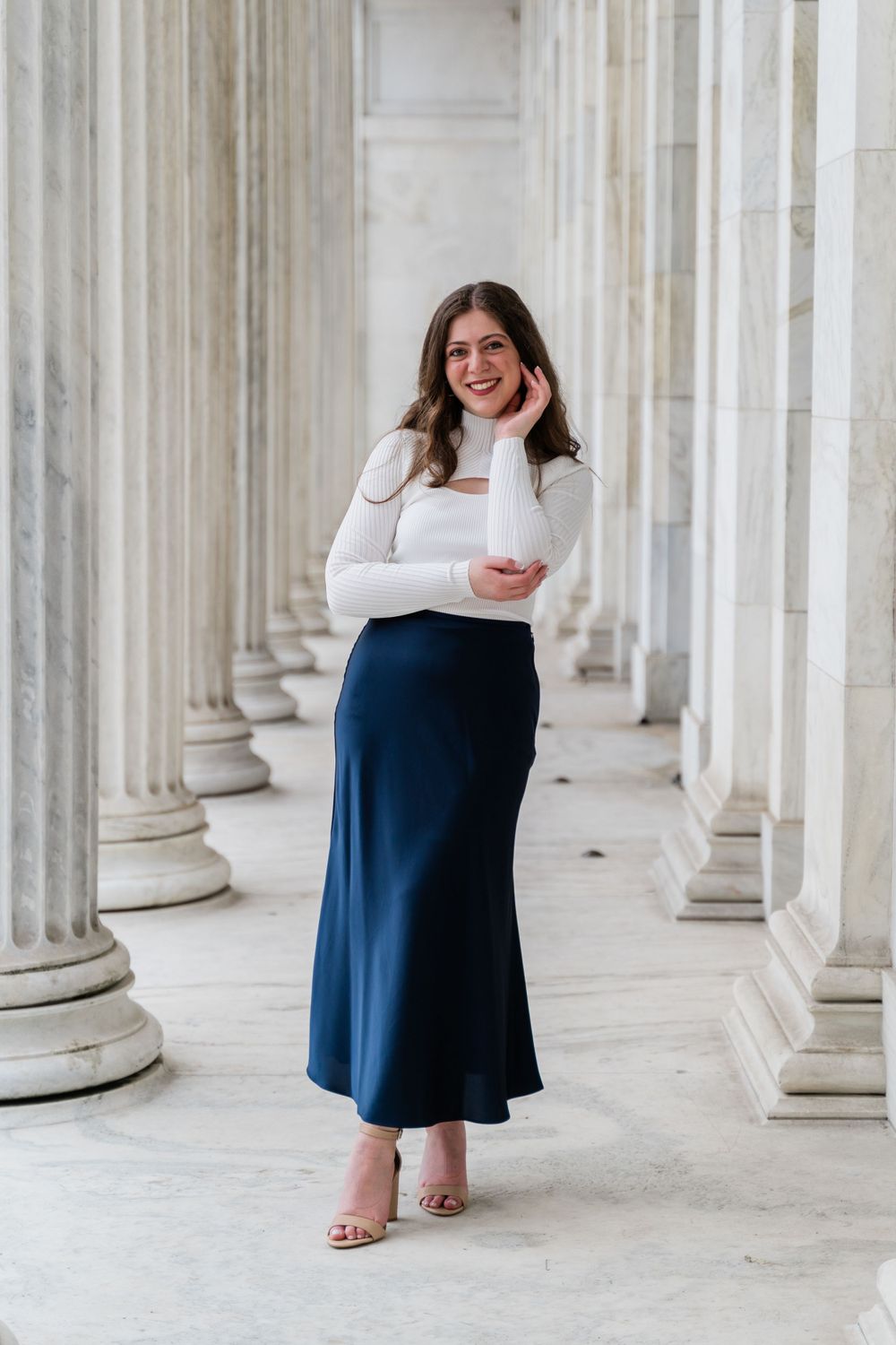 Elegant portrait between white columns showing a casual yet sophisticated outfit with a white top and blue skirt.