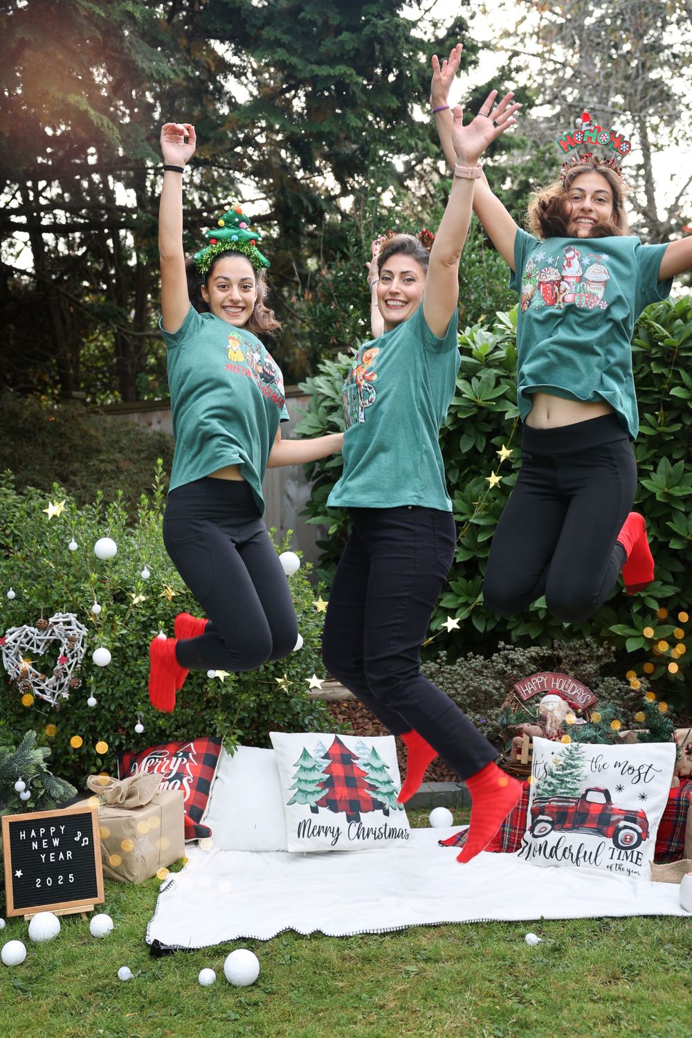 Three friends in matching turquoise shirts jump joyfully in front of a Christmas-themed outdoor photo setup.