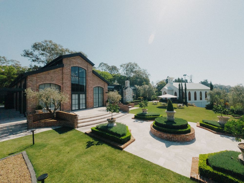 Modern brick wedding venue with manicured gardens and fountain under bright blue sky.
