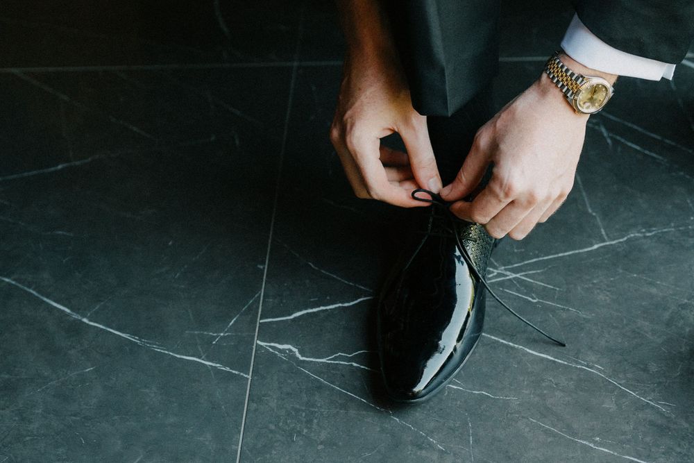 Close-up of hands tying black dress shoe laces on dark marble floor.