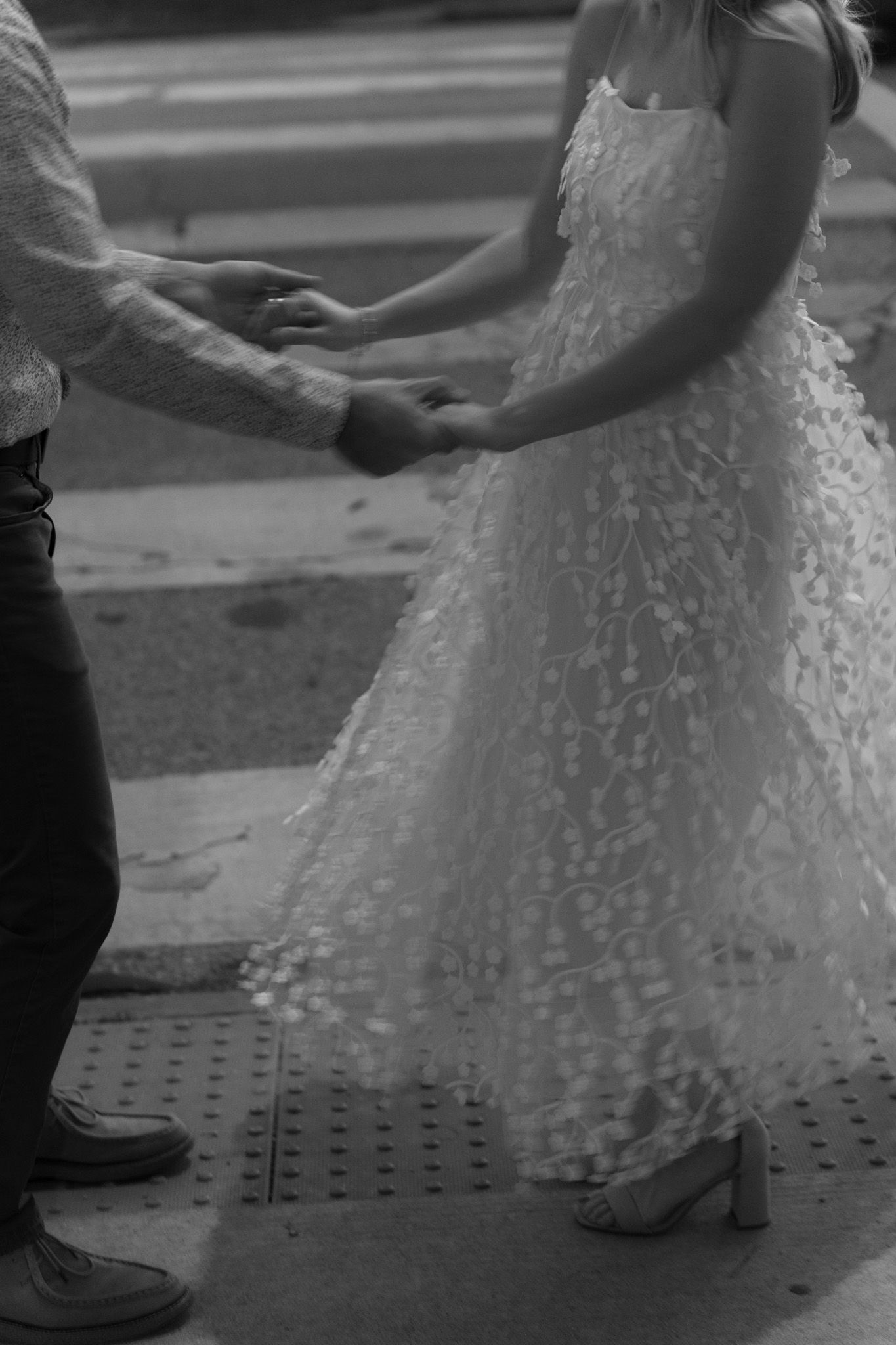 A black and white photo showing hands clasped together with a bride wearing a white lace dress and heels.