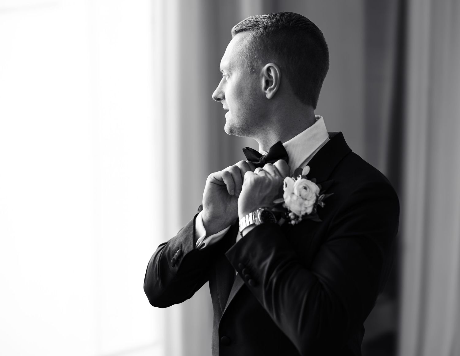Elegant groom in black tuxedo adjusting bow tie by window in black and white photo.