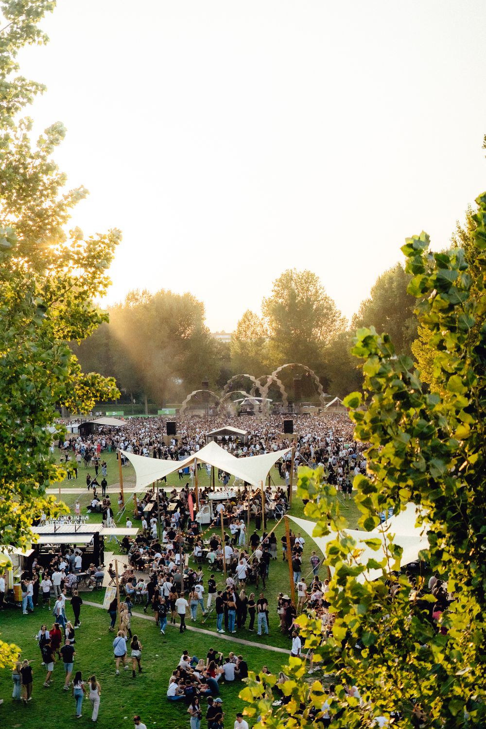Aerial view of large outdoor music festival crowd gathered on grassy field at sunset.