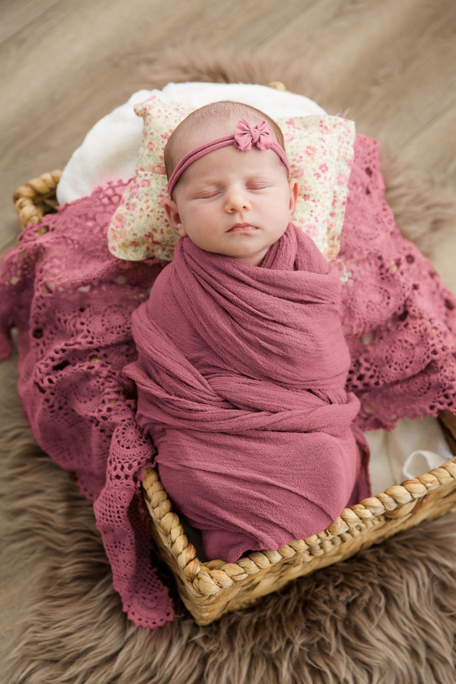 Newborn baby sleeps peacefully wrapped in pink swaddle on lace blanket in woven basket.
