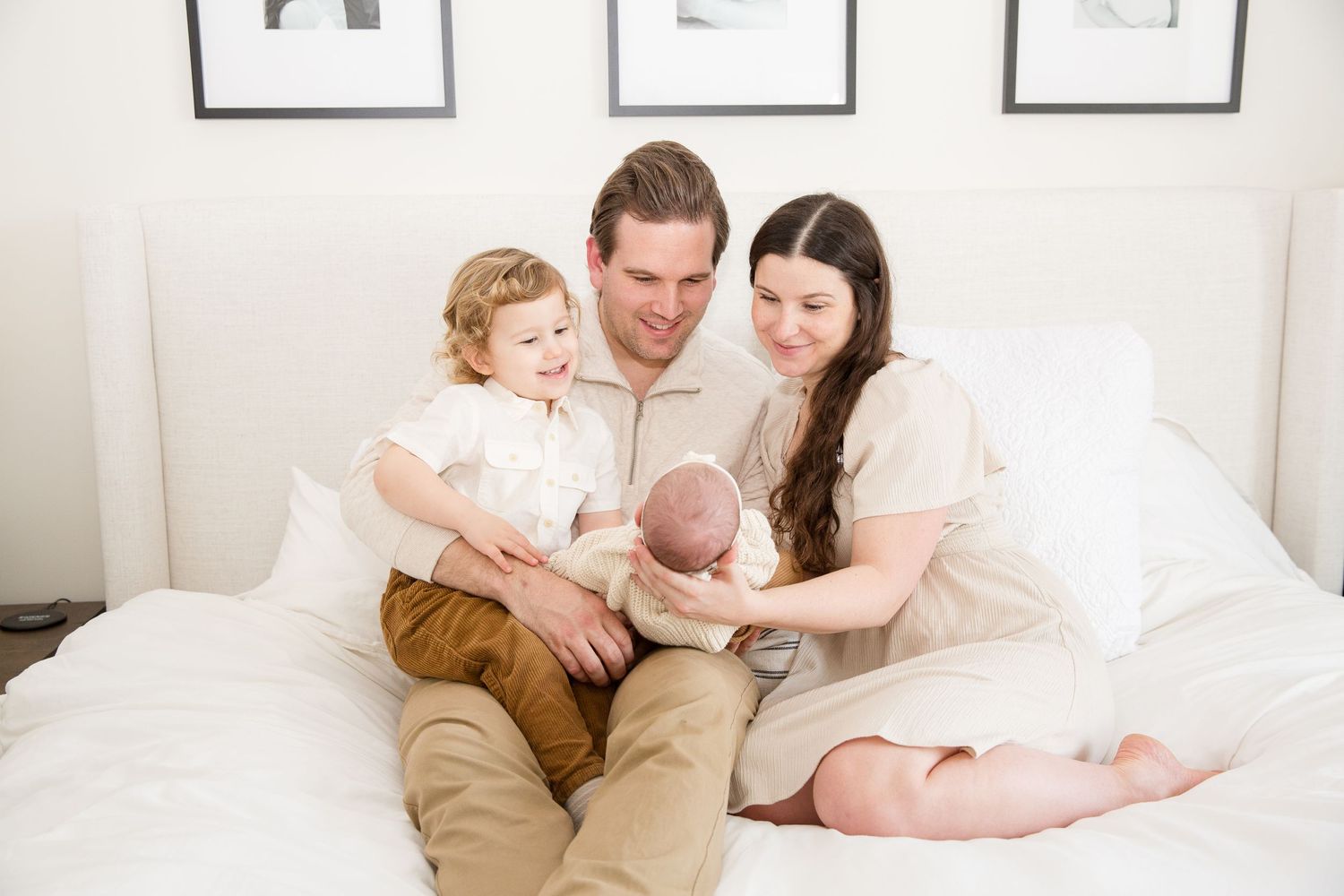 A warm family portrait shows tender moments together on a white bed with framed artwork on the wall.