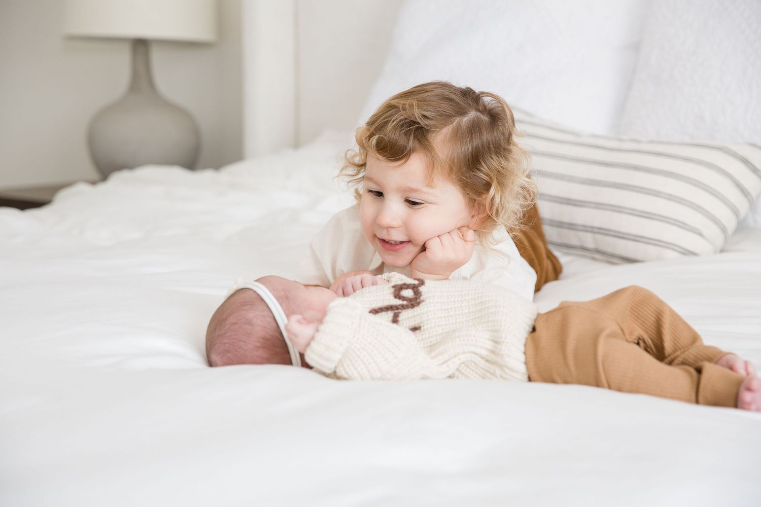 Young child lying on white bedding while looking adoringly at a newborn baby.