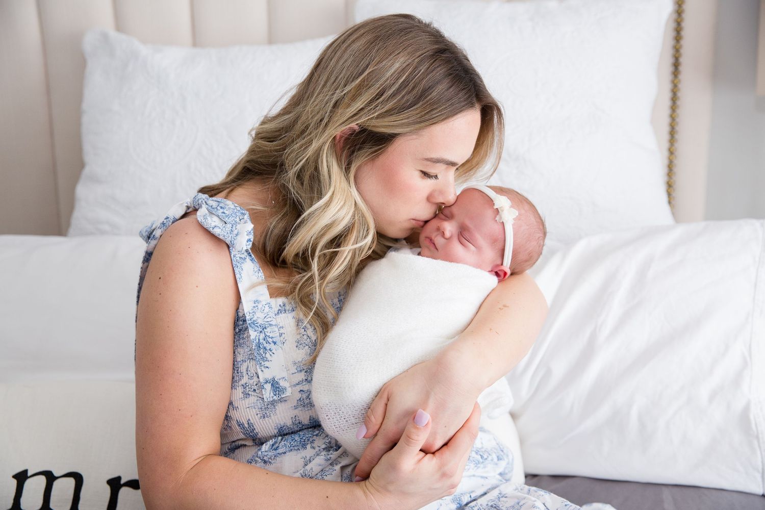 Mother in floral dress tenderly holding newborn baby wrapped in white.
