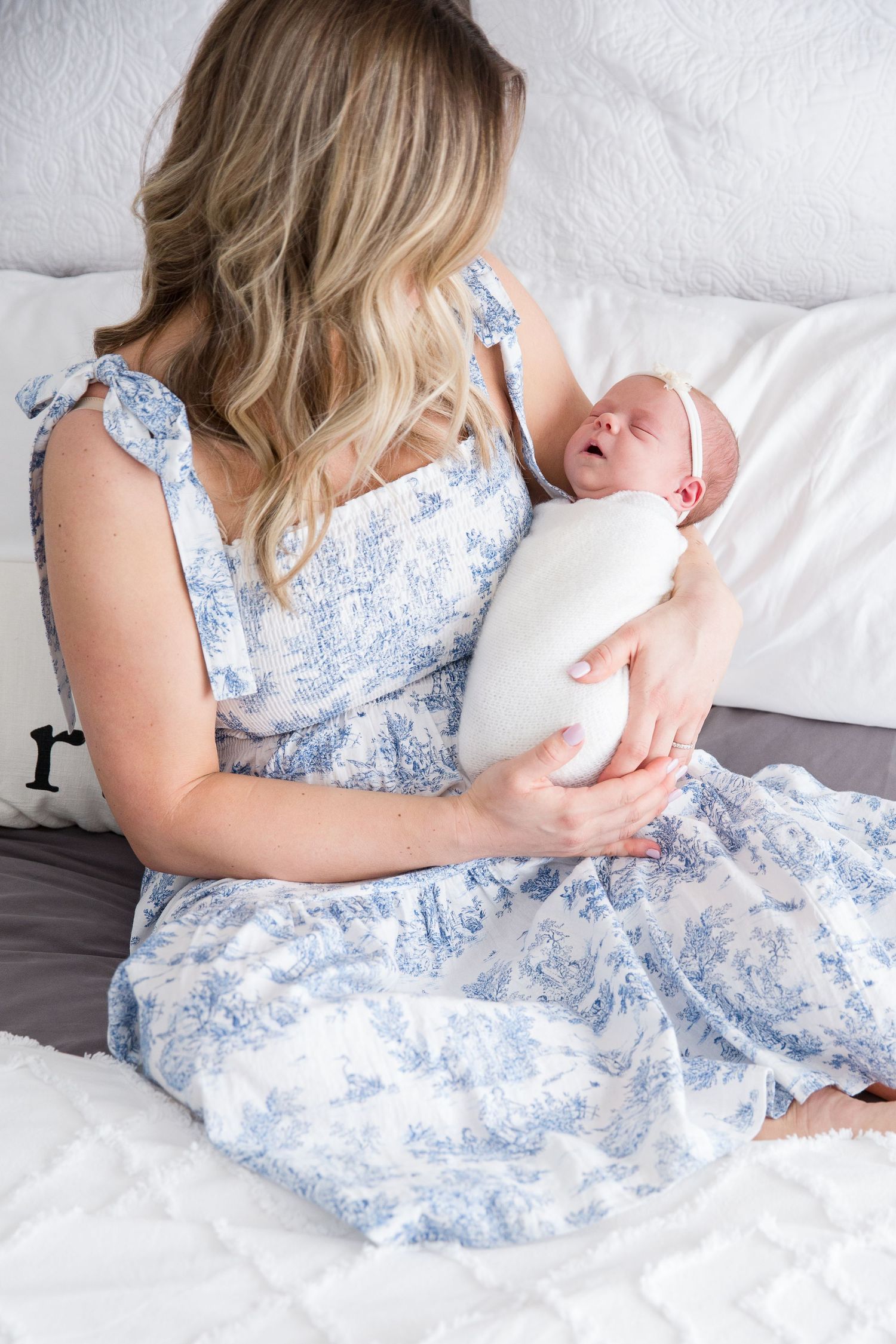 Woman in blue floral dress holding sleeping newborn wrapped in white blanket.