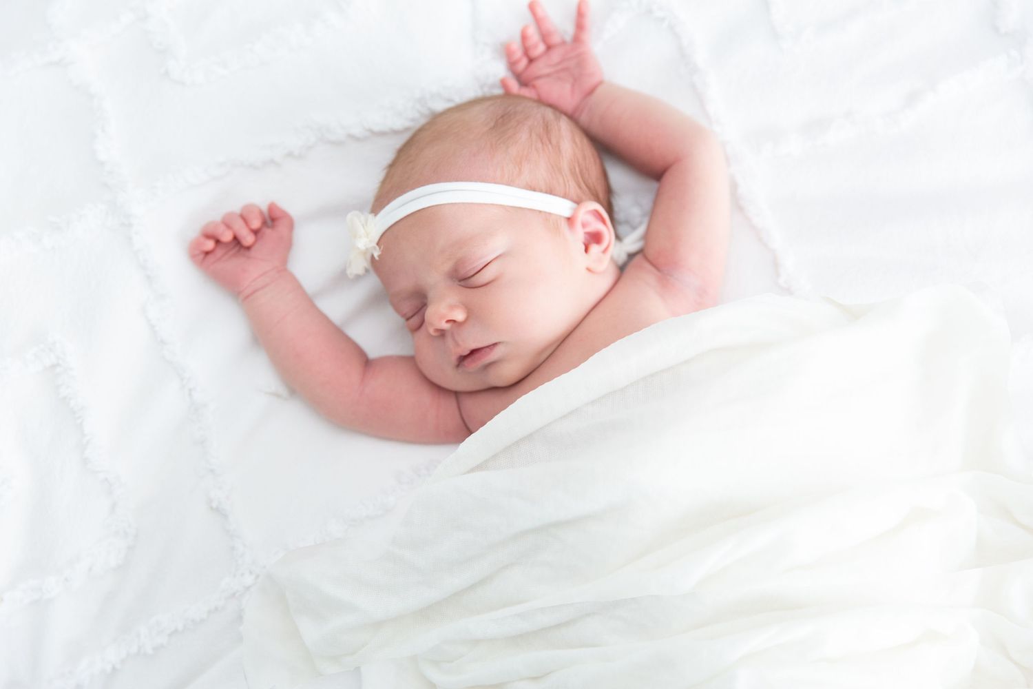 Sleeping newborn baby wrapped in white blanket with arms stretched overhead.