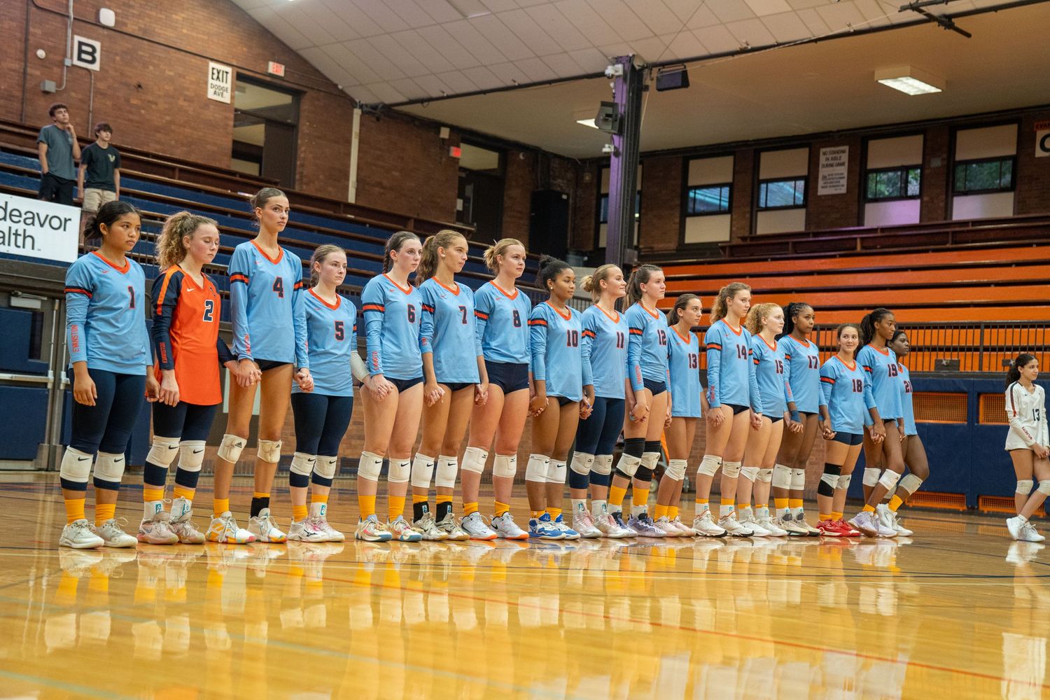Volleyball team in light blue uniforms lined up on court before match.