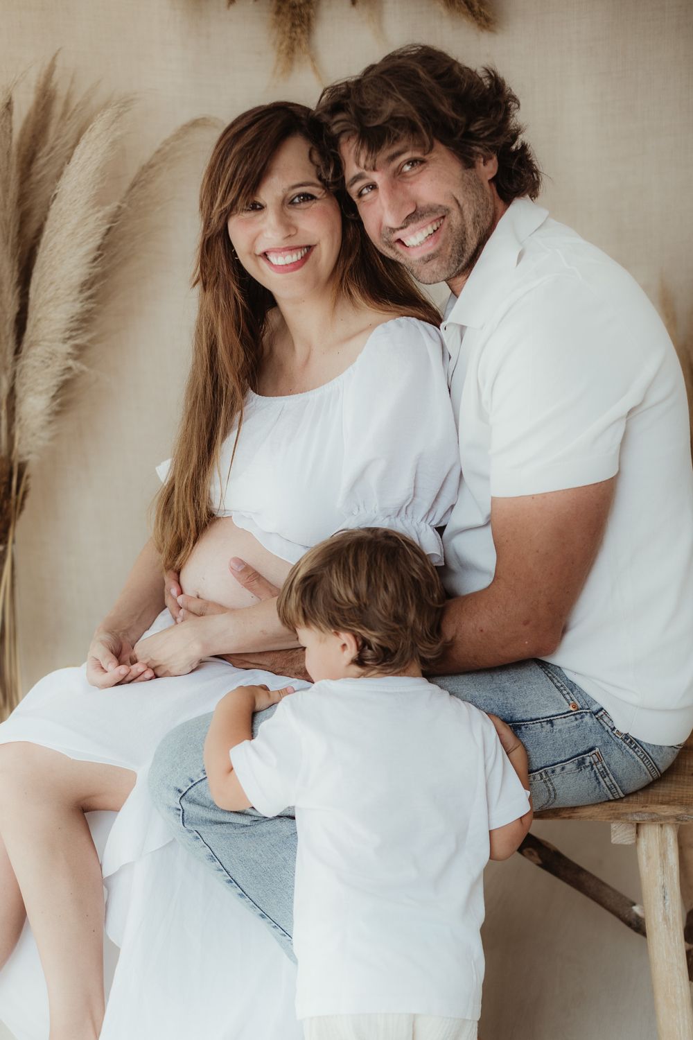 Happy pregnant family with toddler posing in white outfits during maternity photo session with pampas grass backdrop.