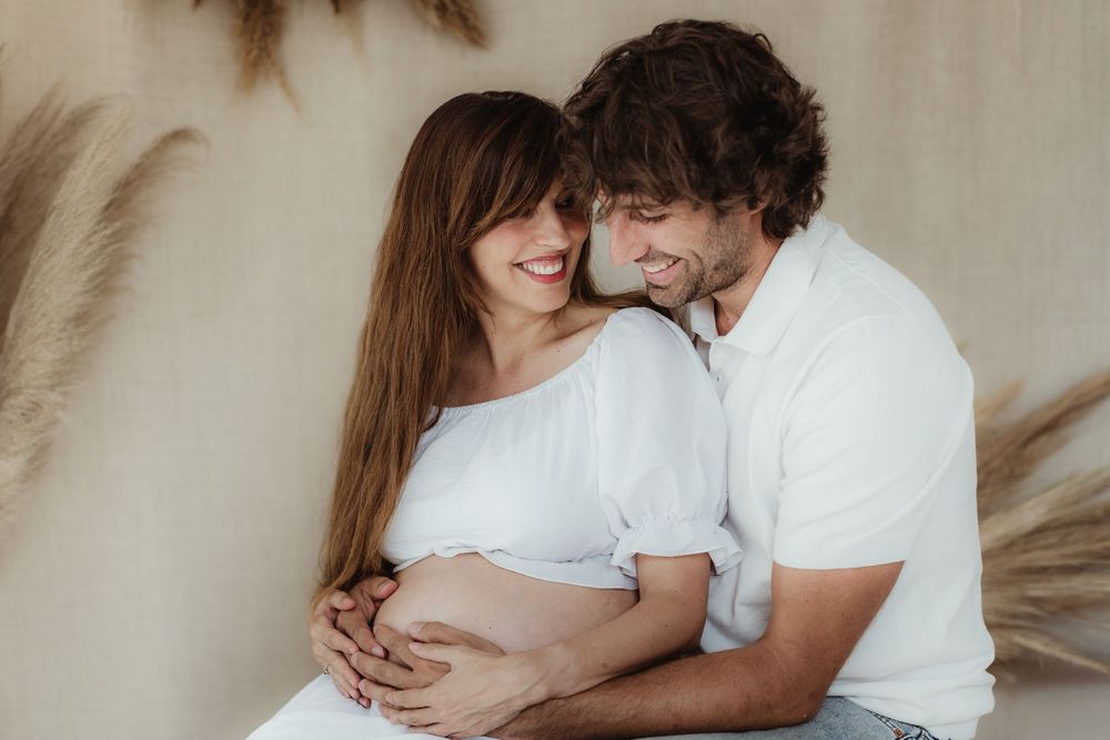 A romantic couple in white outfits embraces intimately during a tender photo session against a neutral background.
