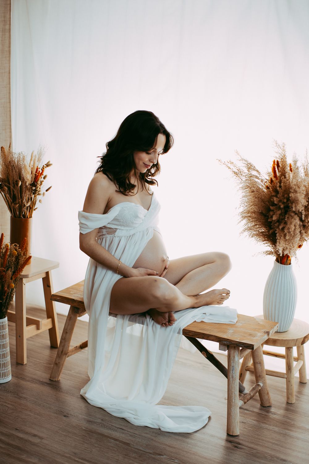 A figure in a flowing white dress poses gracefully with dried pampas grass arrangements in a minimalist indoor setting.