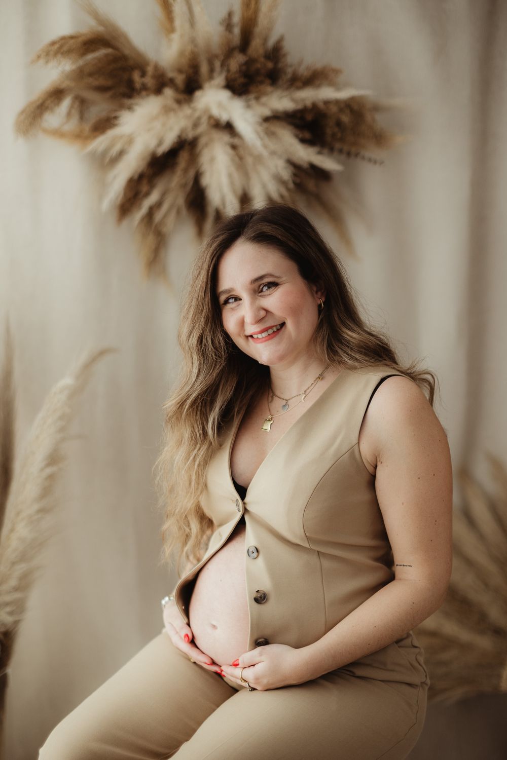 A maternity photo series with pampas grass decor shows a woman in a beige dress posing against a neutral background.