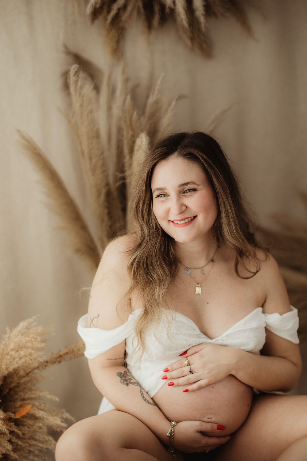 A maternity photo featuring an expectant mother in a white off-shoulder dress posed with dried pampas grass decorations.