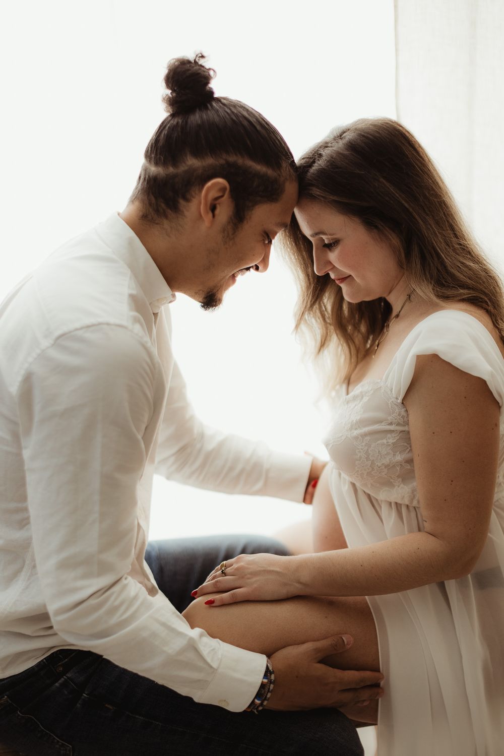 Maternity photo series showing intimate moments between couple in white clothing against bright window light.
