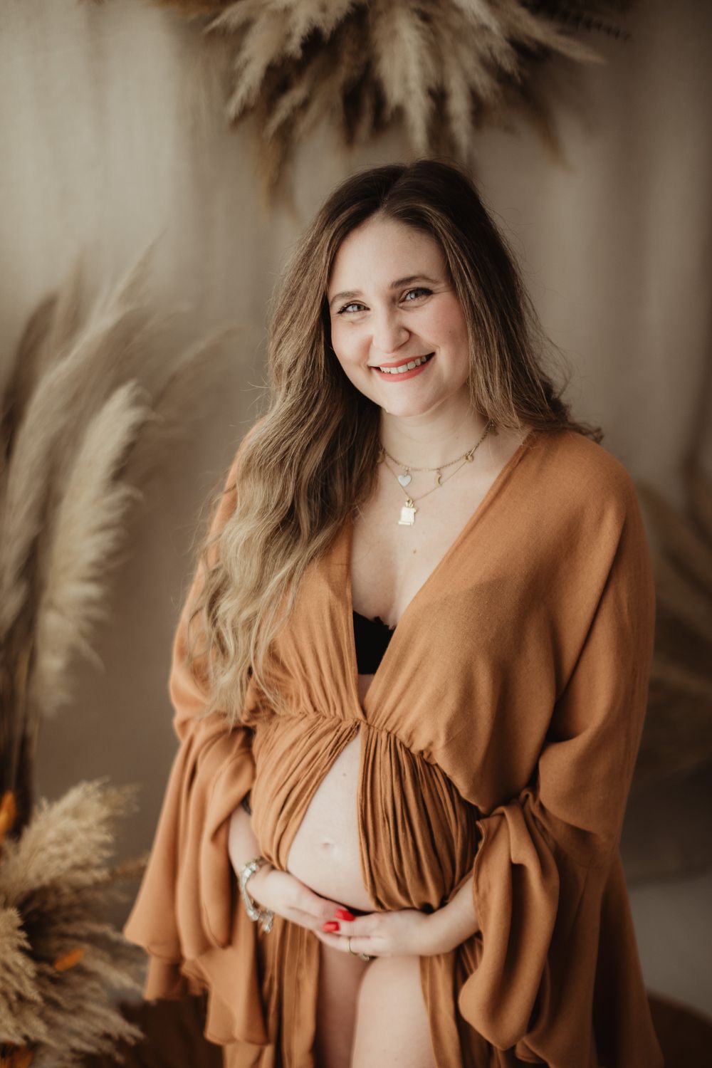 A pregnant woman in a flowing rust-colored dress poses against a neutral backdrop with pampas grass decor.