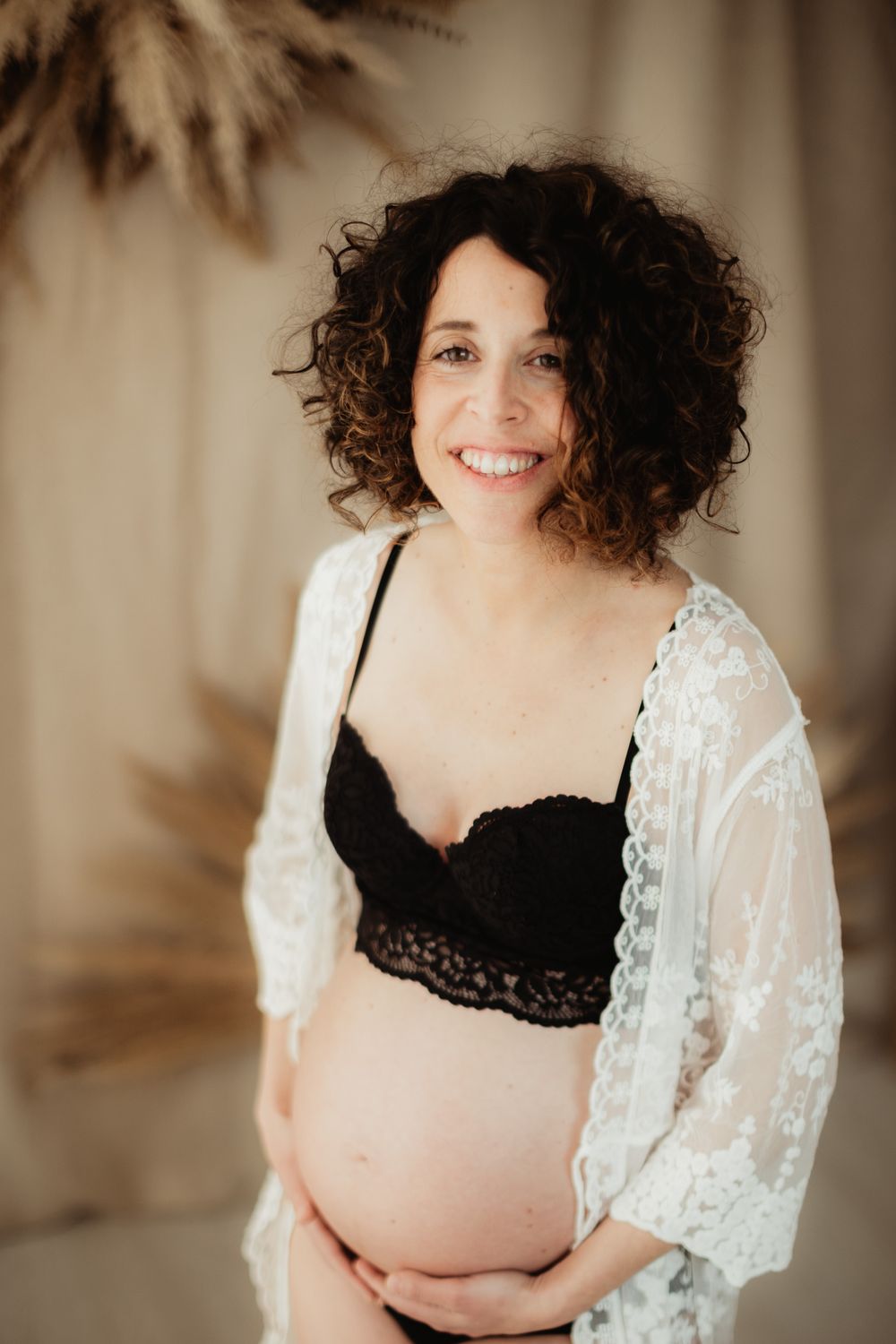 Smiling pregnant woman with curly hair wearing black lace bra and white lace robe, cradling her baby bump.