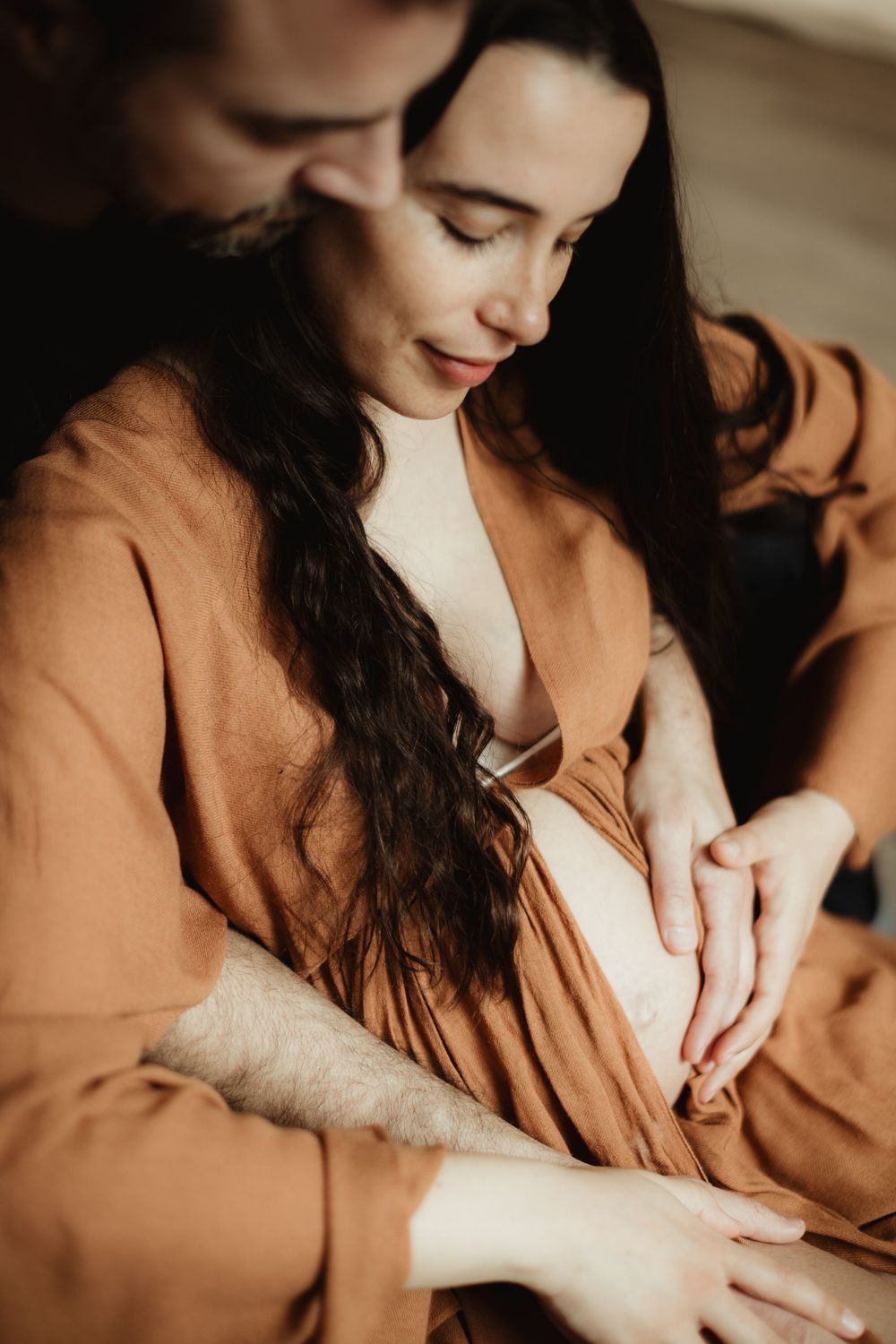 A couple in matching rust-colored robes tenderly cradle a pregnant belly together in an intimate maternity photo.