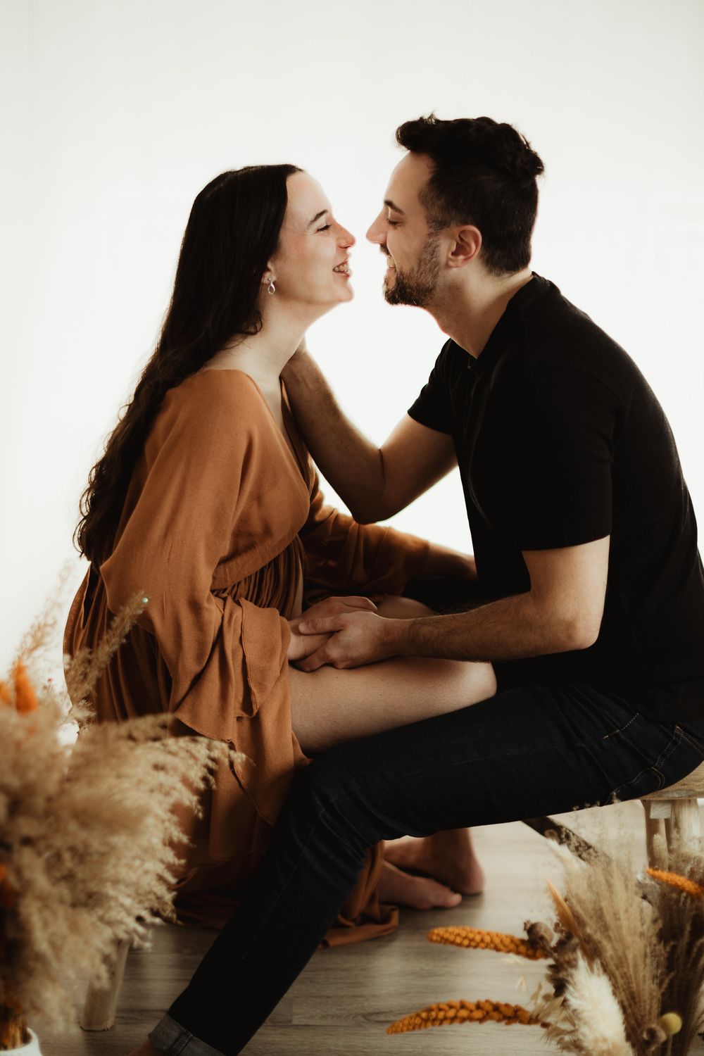A loving couple shares a romantic moment, almost kissing, surrounded by dried pampas grass in a bright studio setting.
