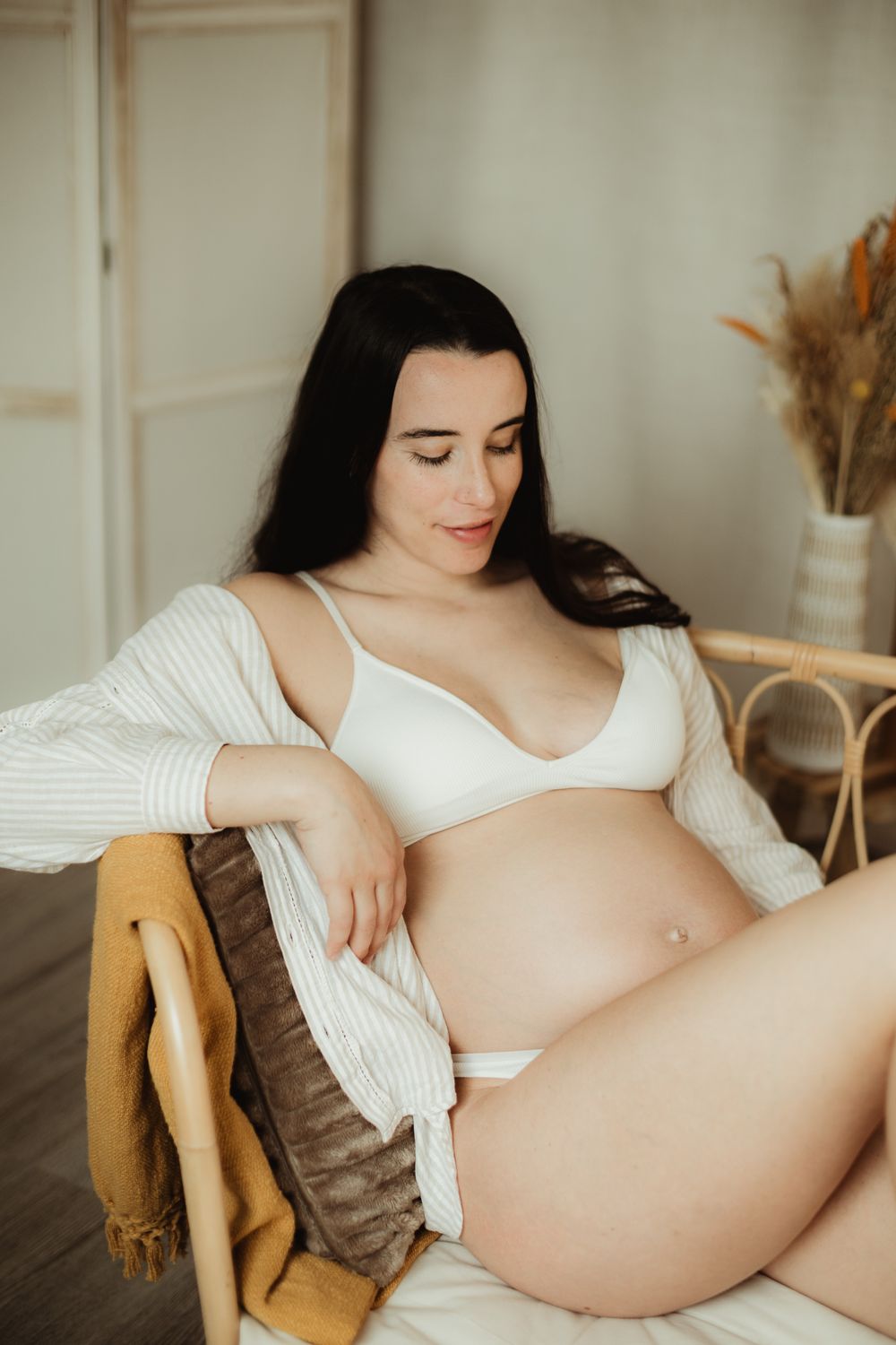 Pregnant woman in white bra and underwear sitting in a rattan chair, gently cradling her baby bump.