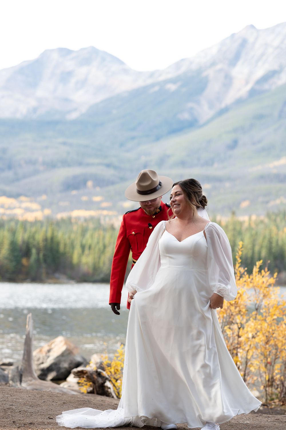 A wedding couple stands on a mountain overlook with a scenic lake and mountain vista in the background.
