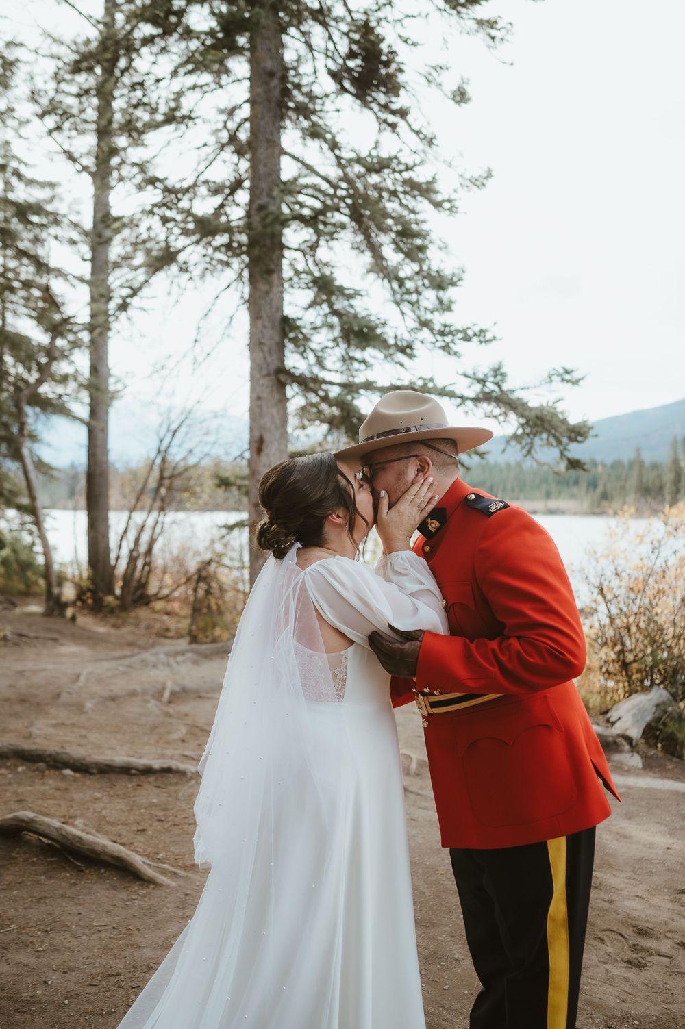 A romantic wedding kiss moment against a backdrop of pine trees and mountain scenery.