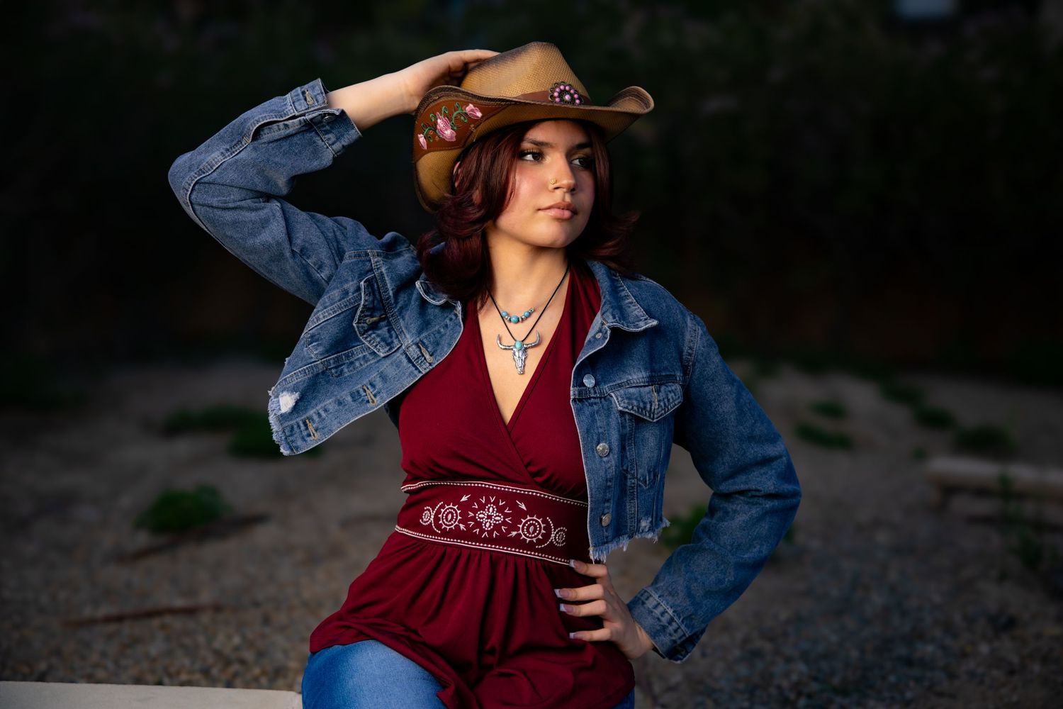 Person in denim jacket and red top poses outdoors at sunset with hand near their brow in artistic portrait.